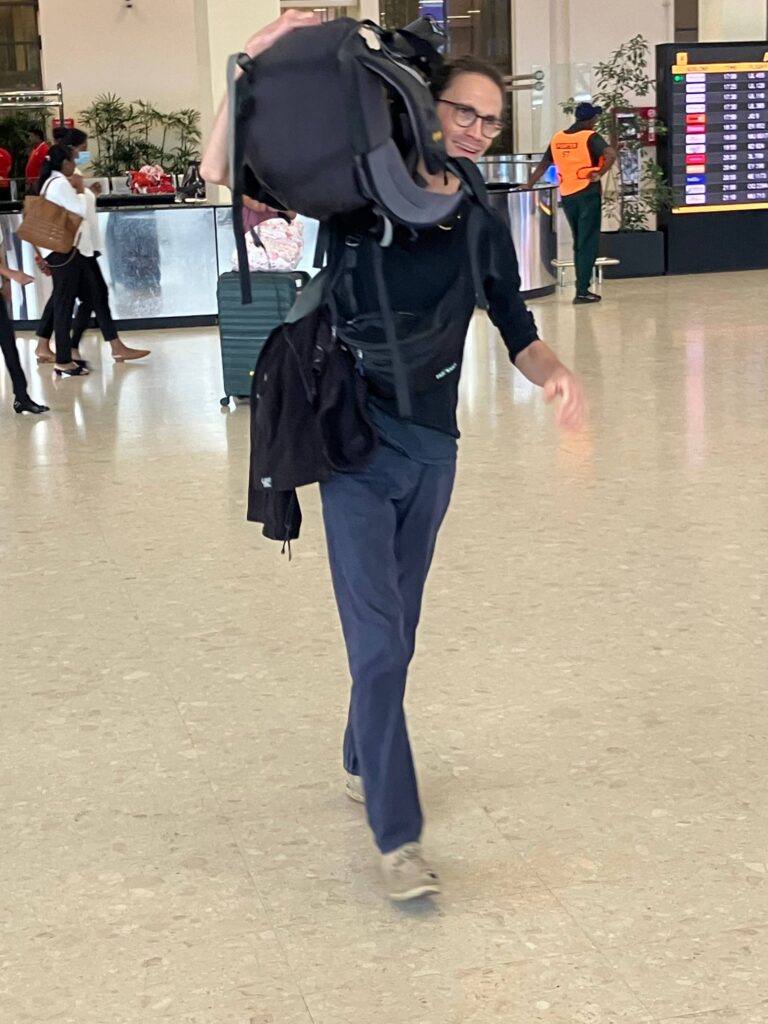 Ein Reisender mit Brille geht in einer Flughafenhalle auf die Kamera zu und trägt einen großen schwarzen Rucksack über der Schulter.
