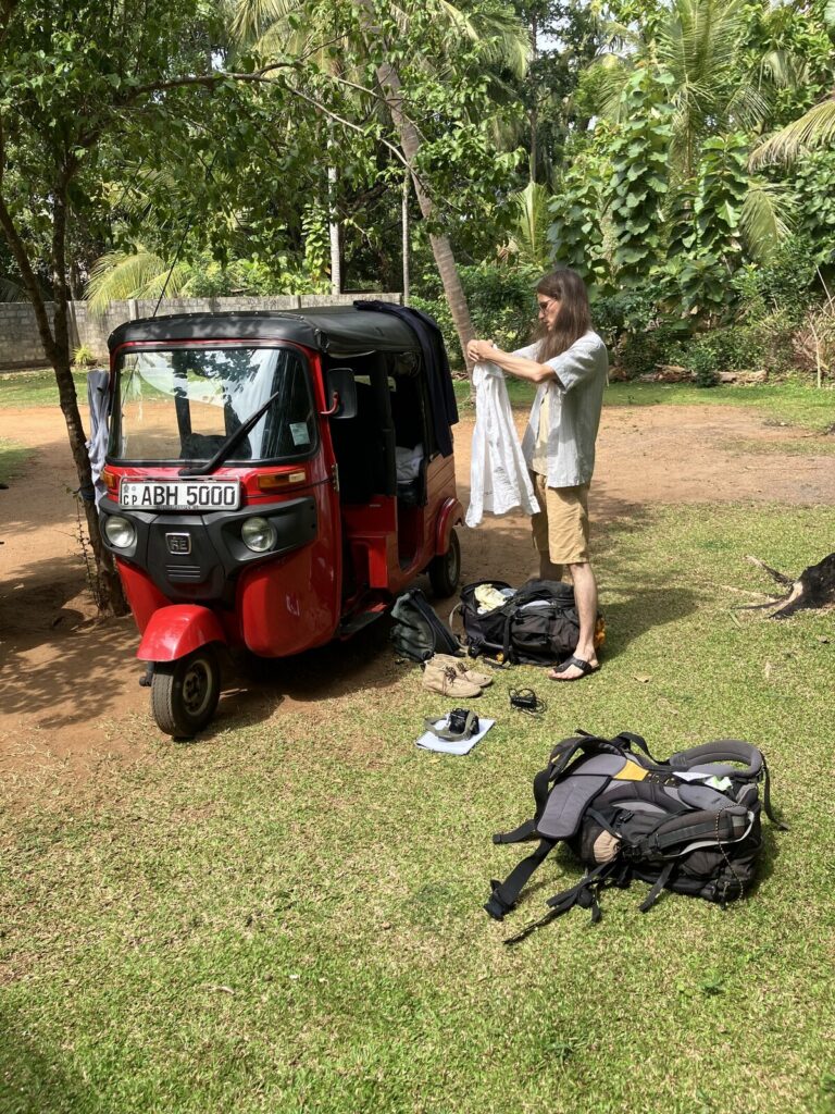 Ein rotes Tuktuk unter Palmen. Daneben steht jemand und faltet ein weißes Hemd; im Gras liegen zwei Rucksäcke, ein Paar Schuhe und eine Kamera auf einem Blatt Papier.