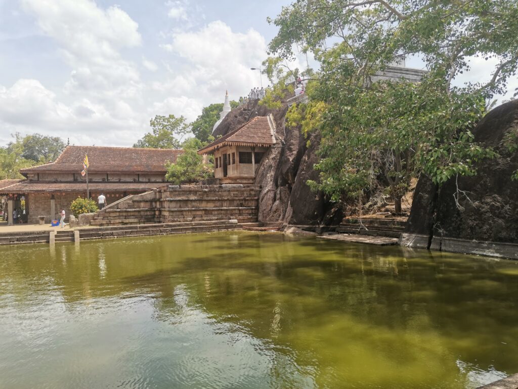 Ein Tempel mit Ziegeldach an einem grünen Teich, dahinter ein großer Felsen mit einem kleinen weißen Stupa und Menschen darauf.