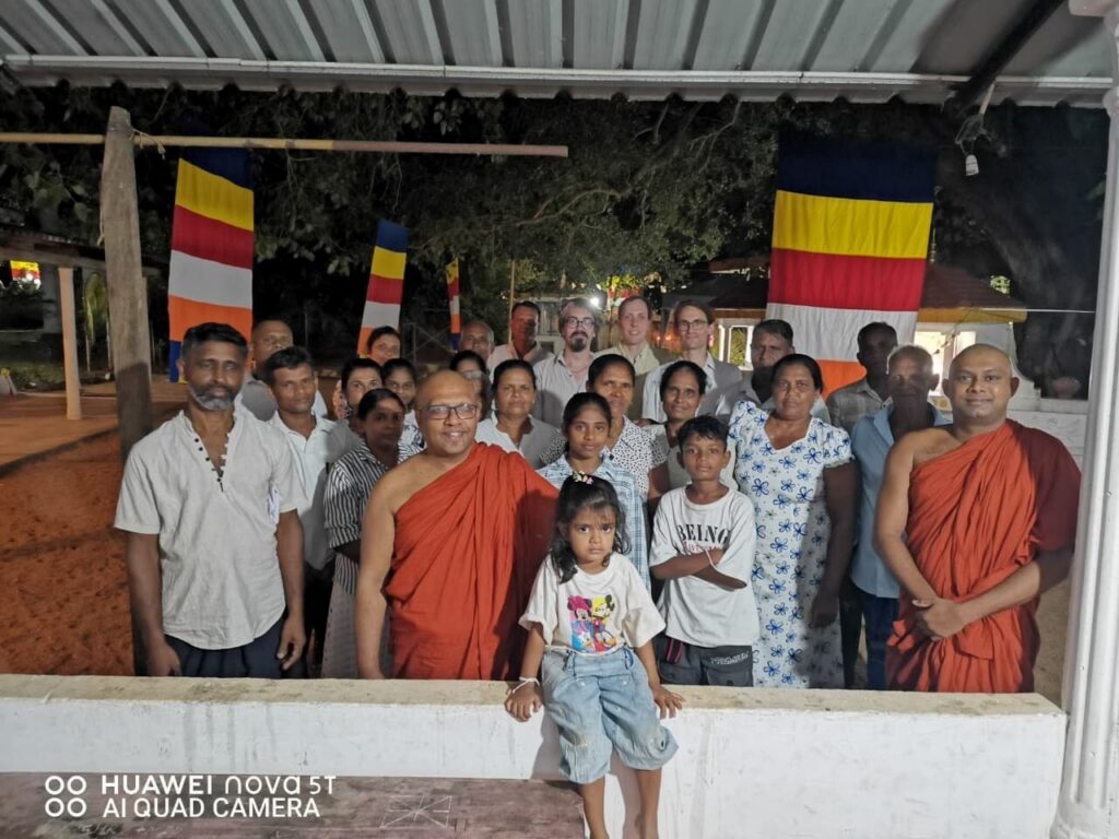 Eine Gruppe von rund zwanzig Dorfbewohnern mit zwei Mönchen in orangen Roben, davor Kinder. In der hinteren Reihe stehen die drei Reisenden.