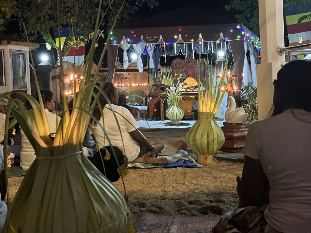 Nachts auf dem Festplatz: Menschen in Weiß sitzen auf dem Boden, im Hintergrund zwei Mönche unter einem Baldachin mit Lichterketten. Vorn stehen von innen beleuchtete Palmblatt-Laternen.