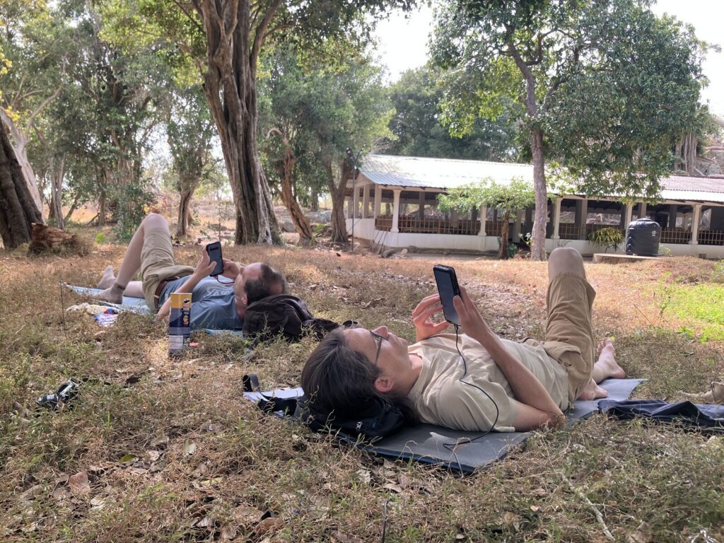 Zwei Menschen liegen auf Isomatten im Schatten unter Bäumen, jeder mit einem Telefon in der Hand. Zwischen ihnen steht eine Dose Chips.