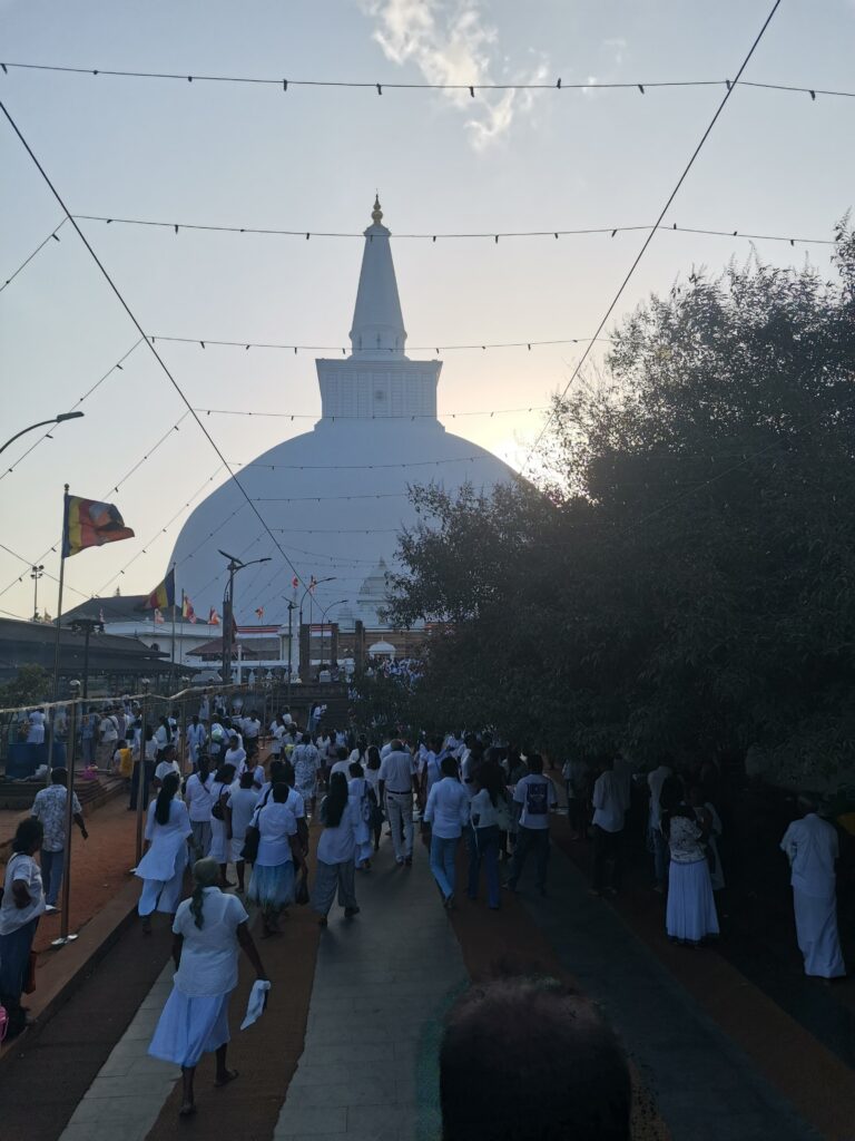 Hunderte Menschen in weißer Kleidung gehen im Abendlicht auf eine große weiße Stupa zu, darüber Lichterketten und Gebetsfahnen.