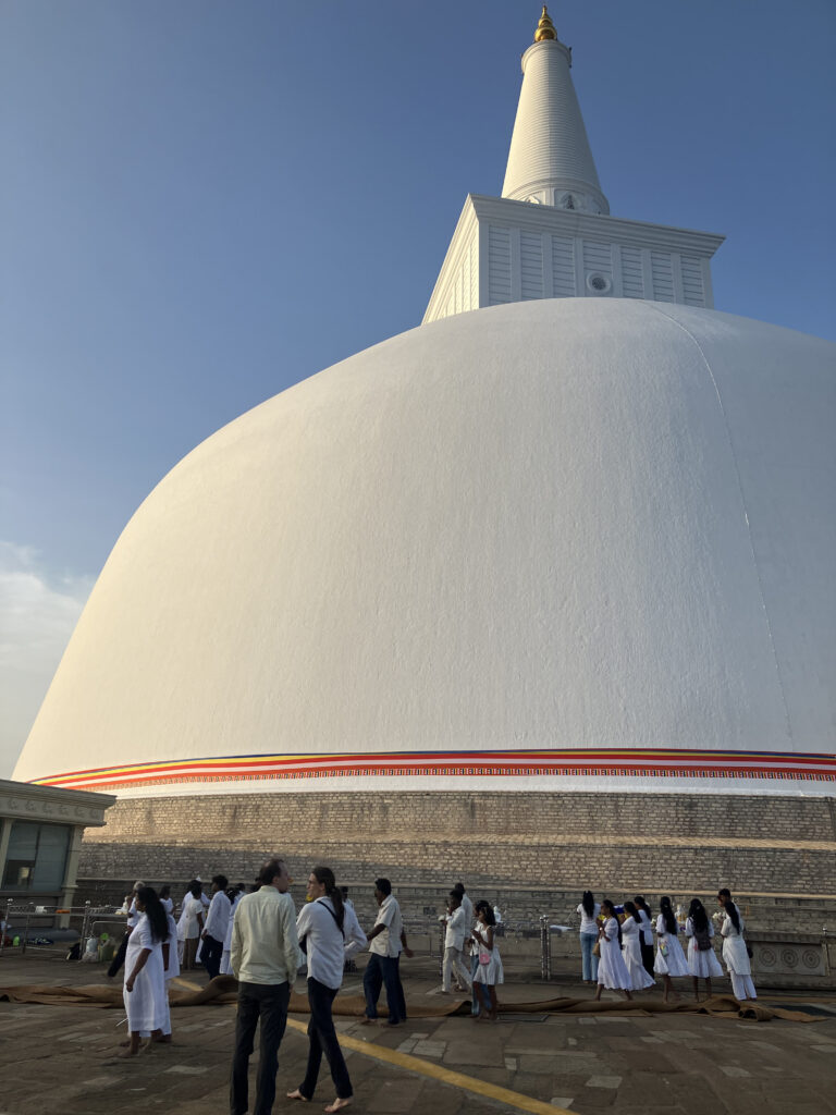 Eine sehr große weiß verputzte Stupa mit goldener Spitze im Abendlicht, davor hunderte Menschen in weißer Kleidung.