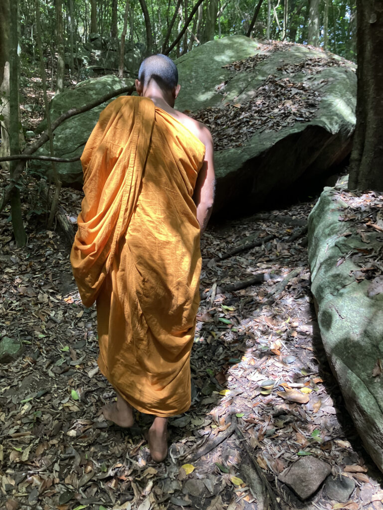 Ein Mönch in oranger Robe geht barfuß auf einem Waldweg zwischen großen, moosbewachsenen Felsblöcken.