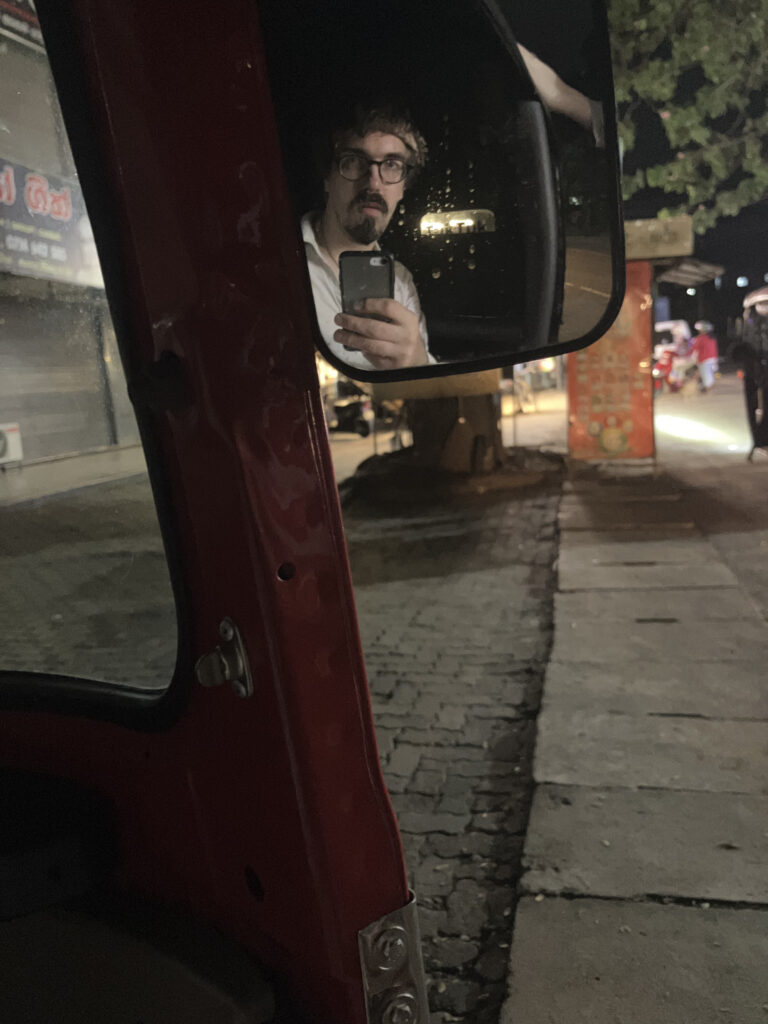 Nächtliche Straßenszene, gespiegelt im Außenspiegel eines TukTuk: ein Mann mit Brille und Bart hält sein Handy hoch, im Hintergrund eine beleuchtete Ladenzeile.