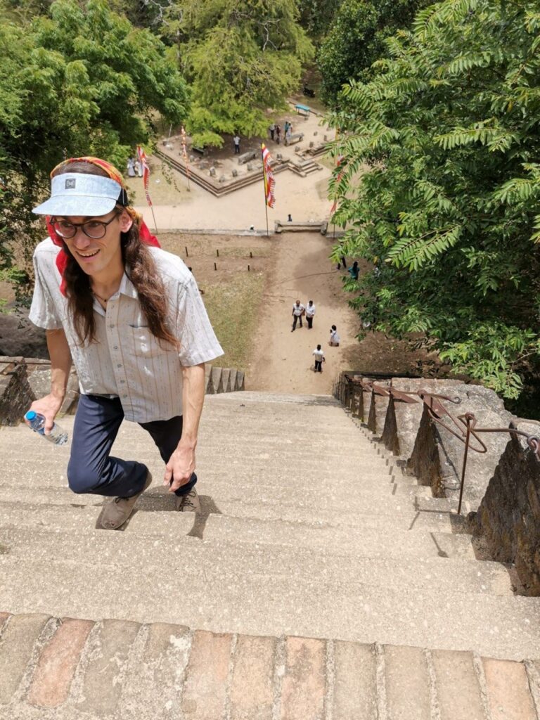 Ein Mann mit langen Haaren, Kopftuch und Sonnenvisier lächelt beim Erklimmen einer sehr steilen Steintreppe, in der Hand eine Wasserflasche.