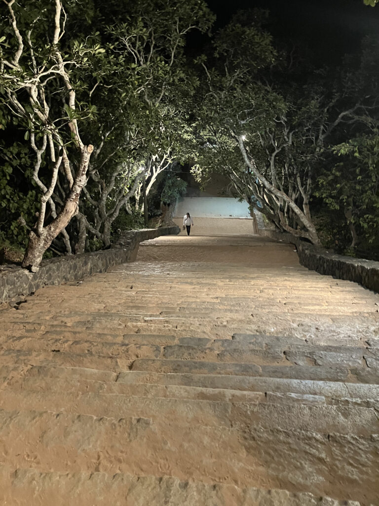 Eine breite, beleuchtete Steintreppe unter Frangipani-Bäumen bei Nacht, oben steht eine einzelne Gestalt.
