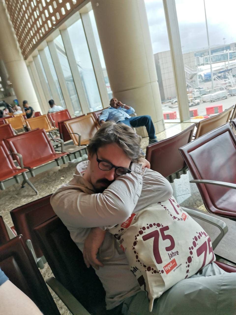 Ein Mann schläft auf einem Sitz in einem Flughafen-Wartebereich und hält eine große Stofftasche im Arm.