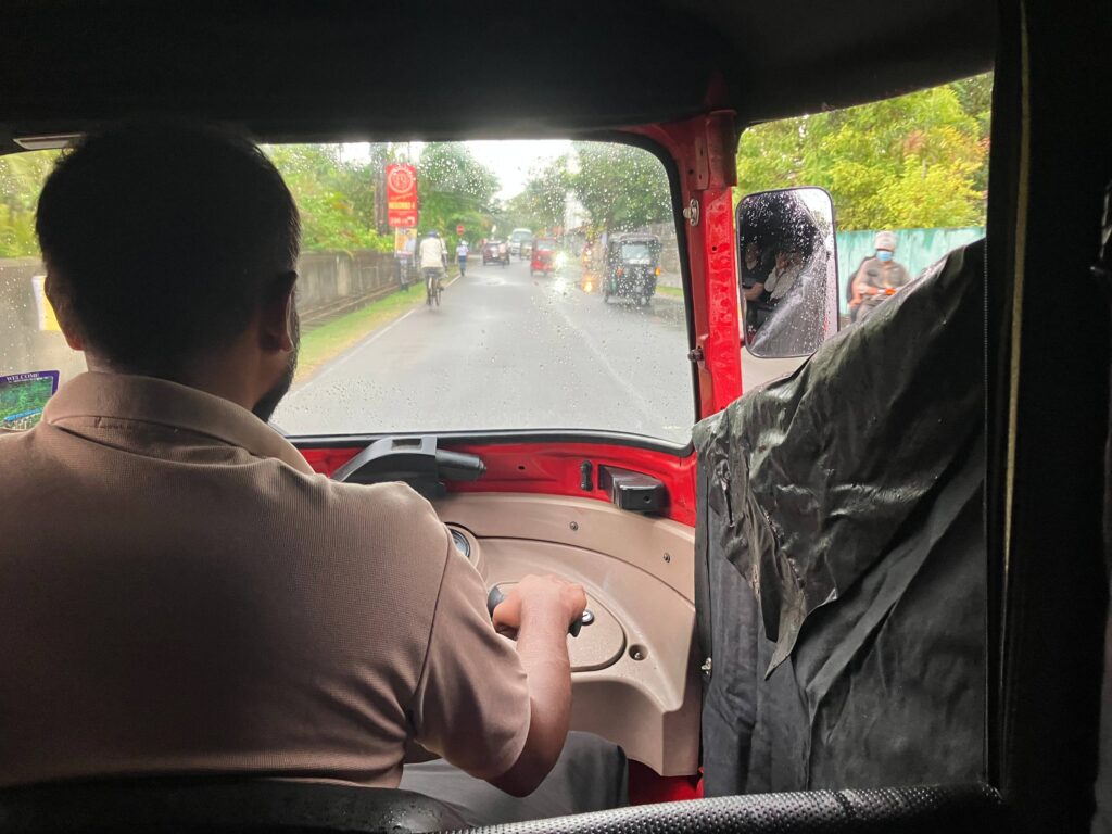 Blick aus dem Fond eines roten Tuk-Tuks durch eine regennasse Frontscheibe auf eine Straße mit Verkehr.