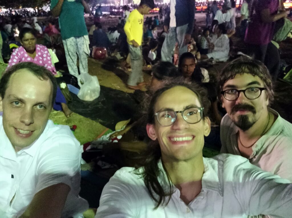 Drei lachende Männer machen ein Selfie, hinter ihnen sitzen viele Menschen auf Planen am Straßenrand.