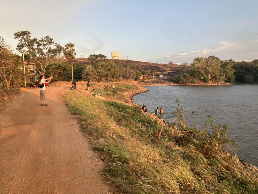 Sonnenuntergang über einem Stausee, Menschen baden im Wasser, auf einem kahlen Hügel steht ein Wasserturm.