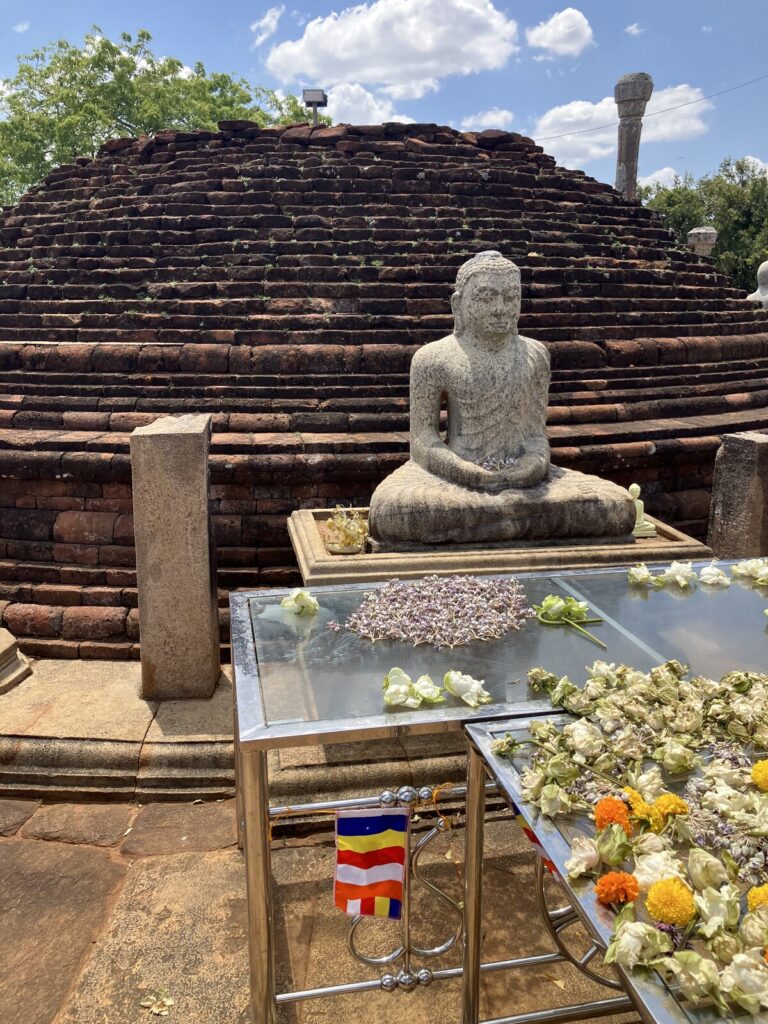 Ein Dagoba aus dunkelrotem, teils bemoostem Backstein, aus den Fugen wächst Gras. Davor eine granitgraue Buddhastatue mit frischen Blüten im Schoß, im Vordergrund zwei Opfertische aus Edelstahl, dicht belegt mit lila-weißen Blüten, Lotosknospen und Ringelblumen. Oben rechts sitzt ein Solarmodul auf einem Mast.