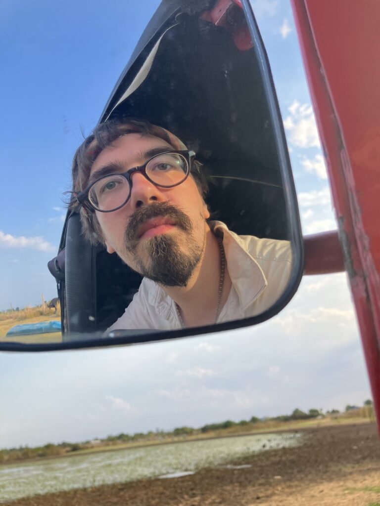 Das Gesicht eines Mannes mit Vollbart und runder dunkler Brille im Rückspiegel eines Tuktuks, von unten aufgenommen. Der rote Rahmen des Fahrzeugs schneidet rechts ins Bild. Am Rand ein blaues Boot am Ufer und die dunkle Silhouette eines gehörnten Tieres auf trockenem Grund.