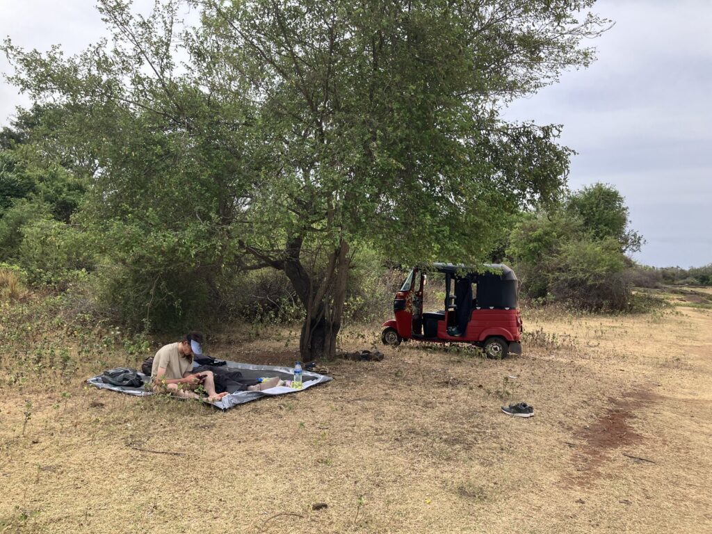 Lagerplatz unter einem einzeln stehenden Baum: eine silbergraue Plane im trockenen Gras, darauf sitzt jemand mit Basecap nach vorn gebeugt. Rechts das rot-schwarze Tuktuk, davor ein Paar Schuhe — und rund zehn Meter weiter, mitten auf der kahlen Erde, ein zweites Paar. Ringsum Dornbuschland.