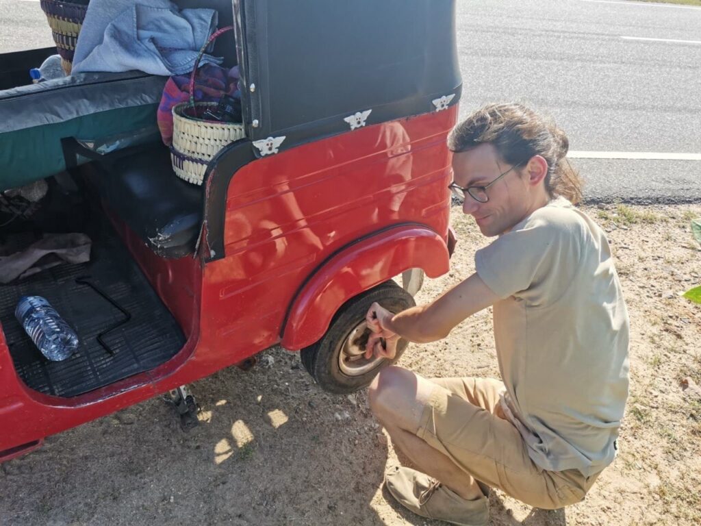 Ein Mann mit langen Haaren hockt mit Werkzeug an einem Rad des TukTuk und grinst schief in die Kamera.