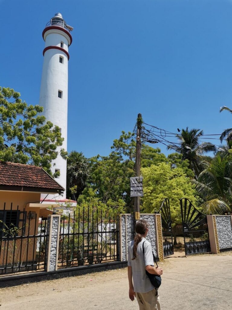 Ein weißer Leuchtturm mit roten Ringen, davor steht ein Mann von hinten.