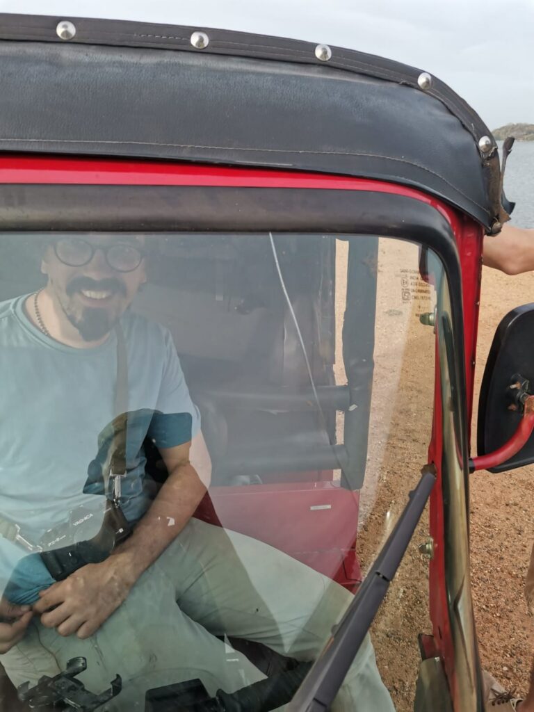Blick durch die Windschutzscheibe eines Tuktuks: Auf dem Fahrersitz sitzt ein Mann mit Vollbart und runder Brille und grinst in die Kamera. Quer über seinem Schoß liegt eine analoge Spiegelreflexkamera. Auf dem Dach liegt der Unterarm einer zweiten Person.