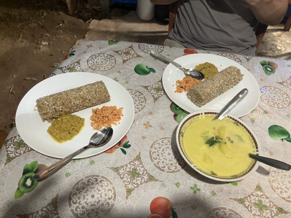Blick von oben auf einen Tisch mit Wachstuchdecke im Obstmuster. Zwei Teller mit je einer Rolle Pittu, gelbem Dal und einem Häufchen orangerotem Sambol, dazu eine gemeinsame Schüssel mit gelbem Kartoffelcurry und Curryblättern. Am oberen Bildrand der Oberkörper einer Person im grauen T-Shirt.